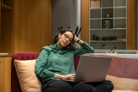 Frau mit Brille sitzt mit skeptischem Blick auf einem roten Sofa und schaut auf einen aufgeklappten Laptop.