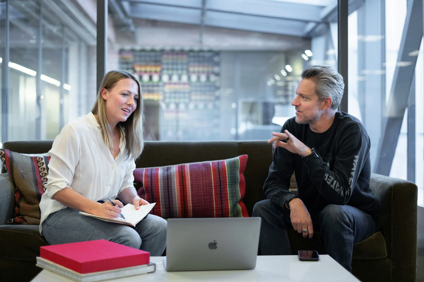 Zwei Personen sitzen auf einem Sofa in einem hellen Büro und unterhalten sich, eine hält ein Notizbuch, die andere deutet auf einen aufgeklappten MacBook.