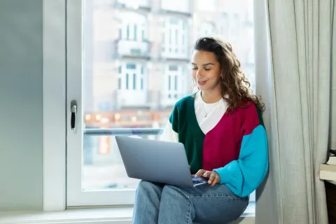 Junge Frau im bunten Strickpullover sitzt lächelnd auf einer Fensterbank und tippt auf einem Laptop.